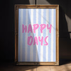 Framed artwork with 'Happy Days' text on a striped background, placed against a dark wall.